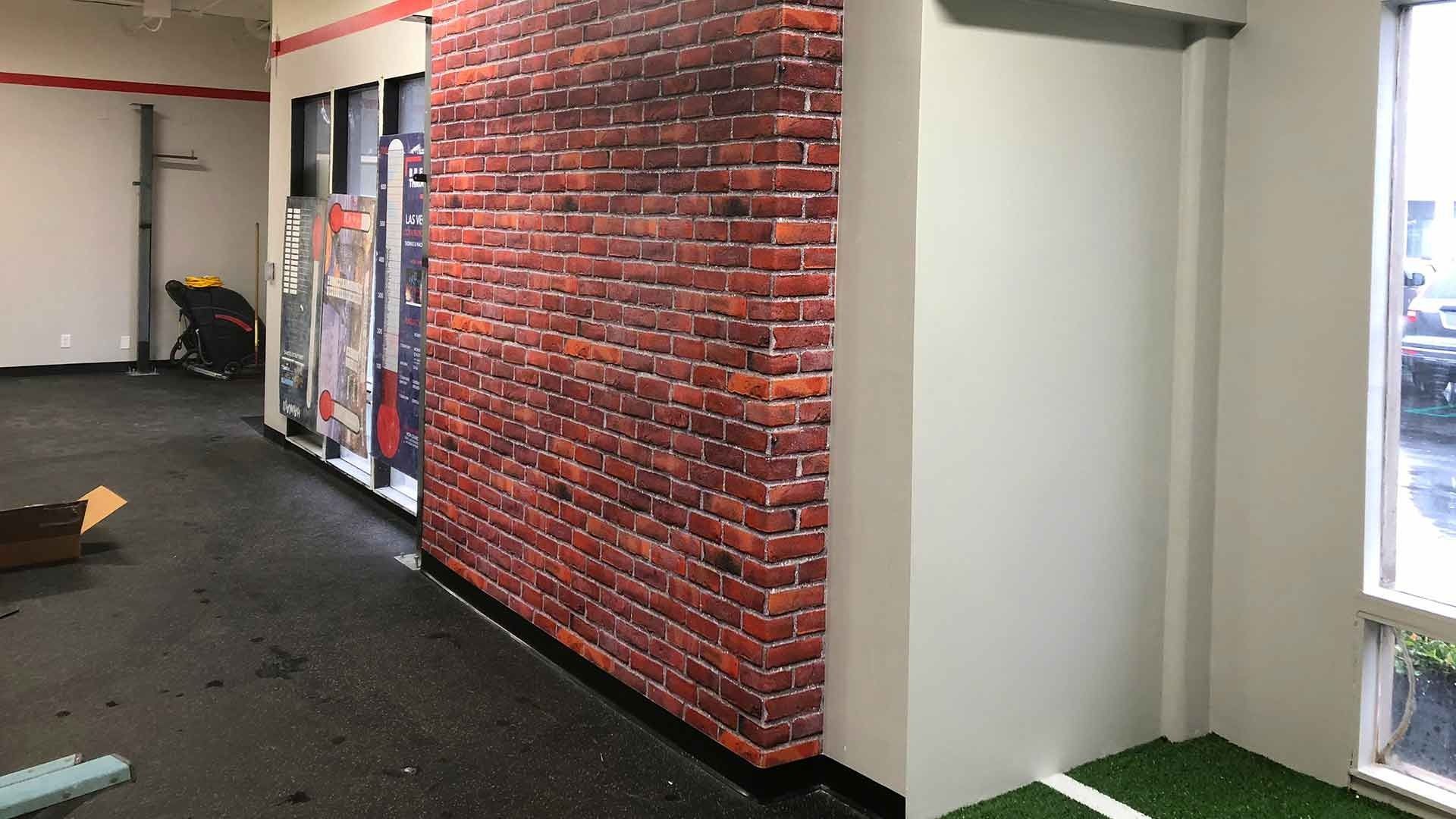 A gym interior with a brick accent wall, black rubber flooring, and a green turf strip near a window.