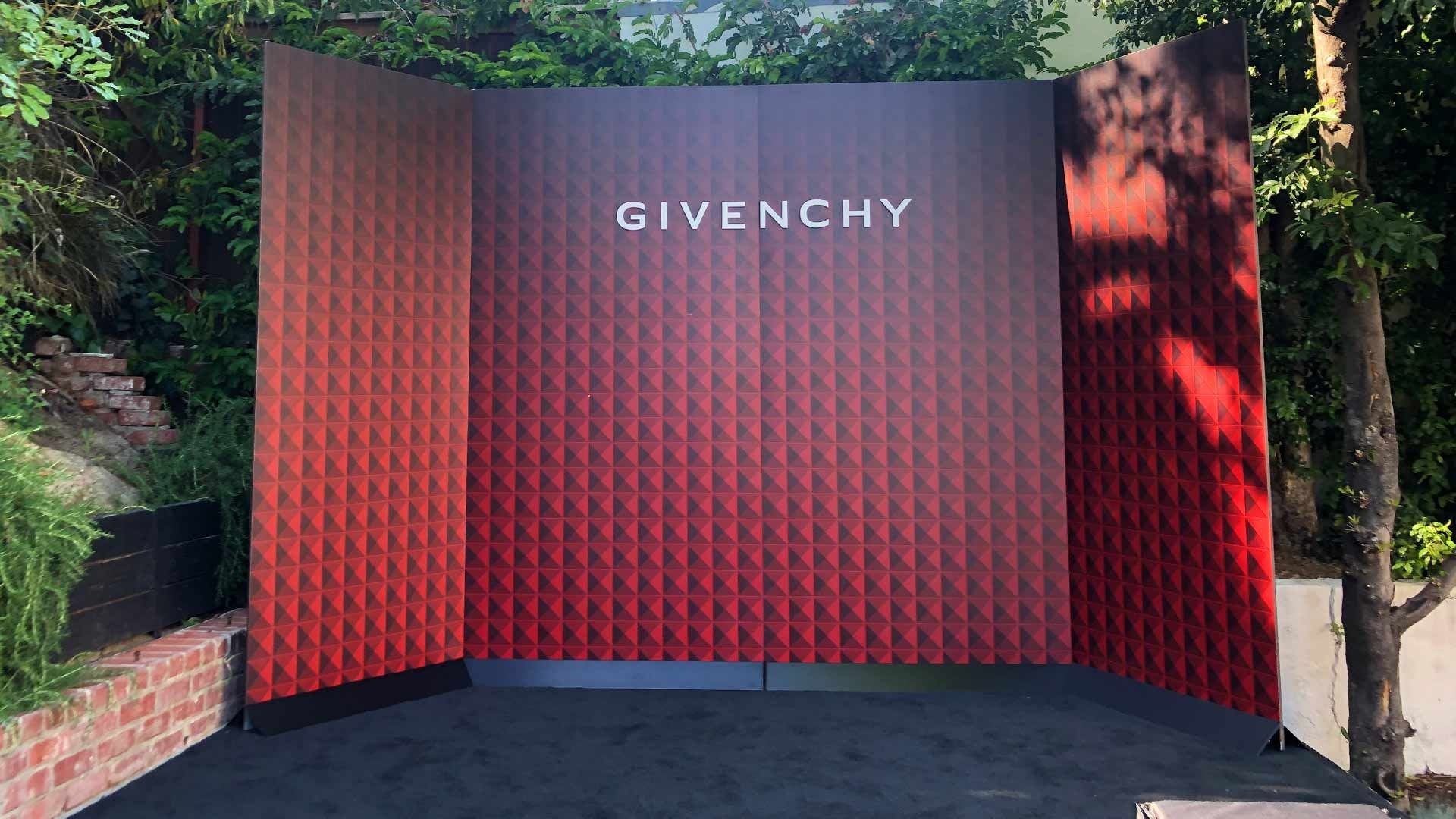 A Givenchy branded backdrop with a red and black geometric gradient pattern, set on a stage outdoors among trees.