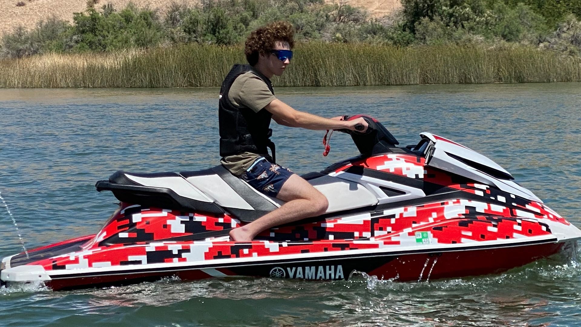 A person in a life vest rides a red, white, and black camouflage-patterned Yamaha jet ski on a sunny, calm lake.