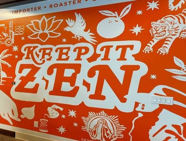 A bright orange wall featuring white line art of a lotus, tiger, coffee brewer, and the text 