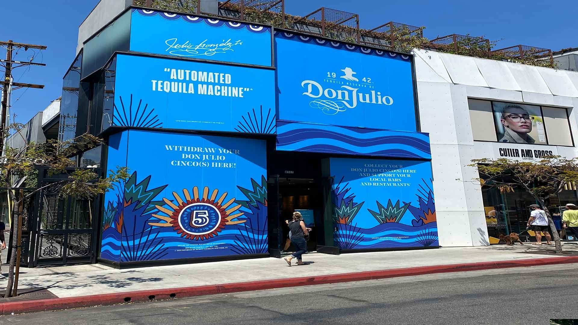 A storefront with bright blue Don Julio branding, a logo, and agave graphics, situated on a sunny street corner.