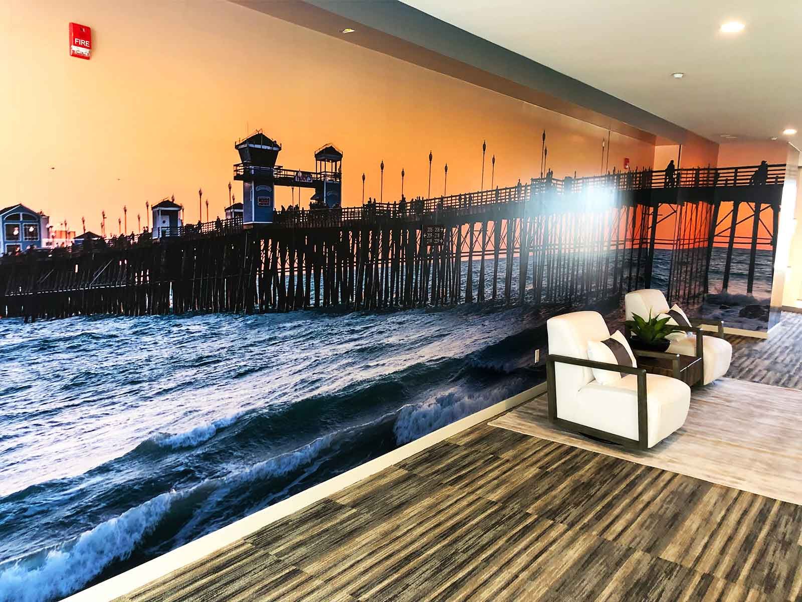 A mural of a pier over a turbulent ocean covers a wall in a lounge with two white armchairs and striped flooring.