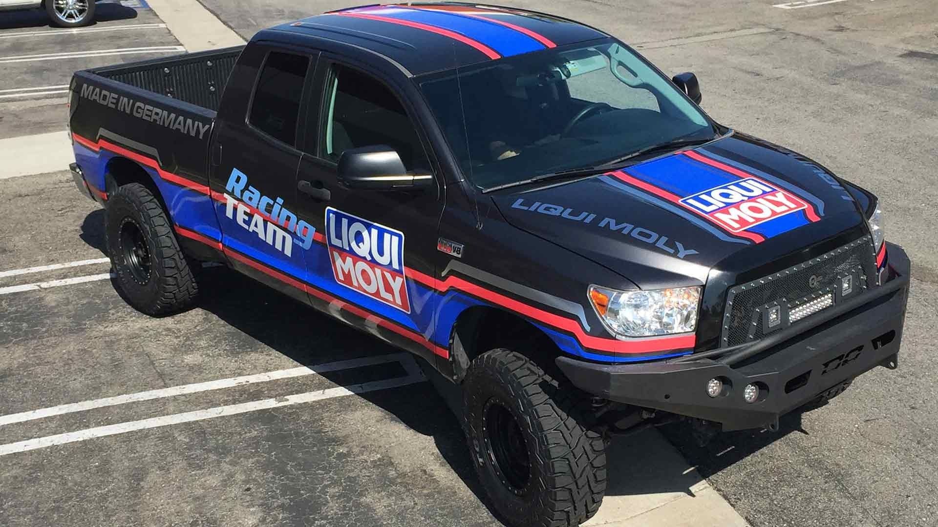 A black pickup truck featuring blue and red racing stripes and Liqui Moly branding parked in an asphalt lot.