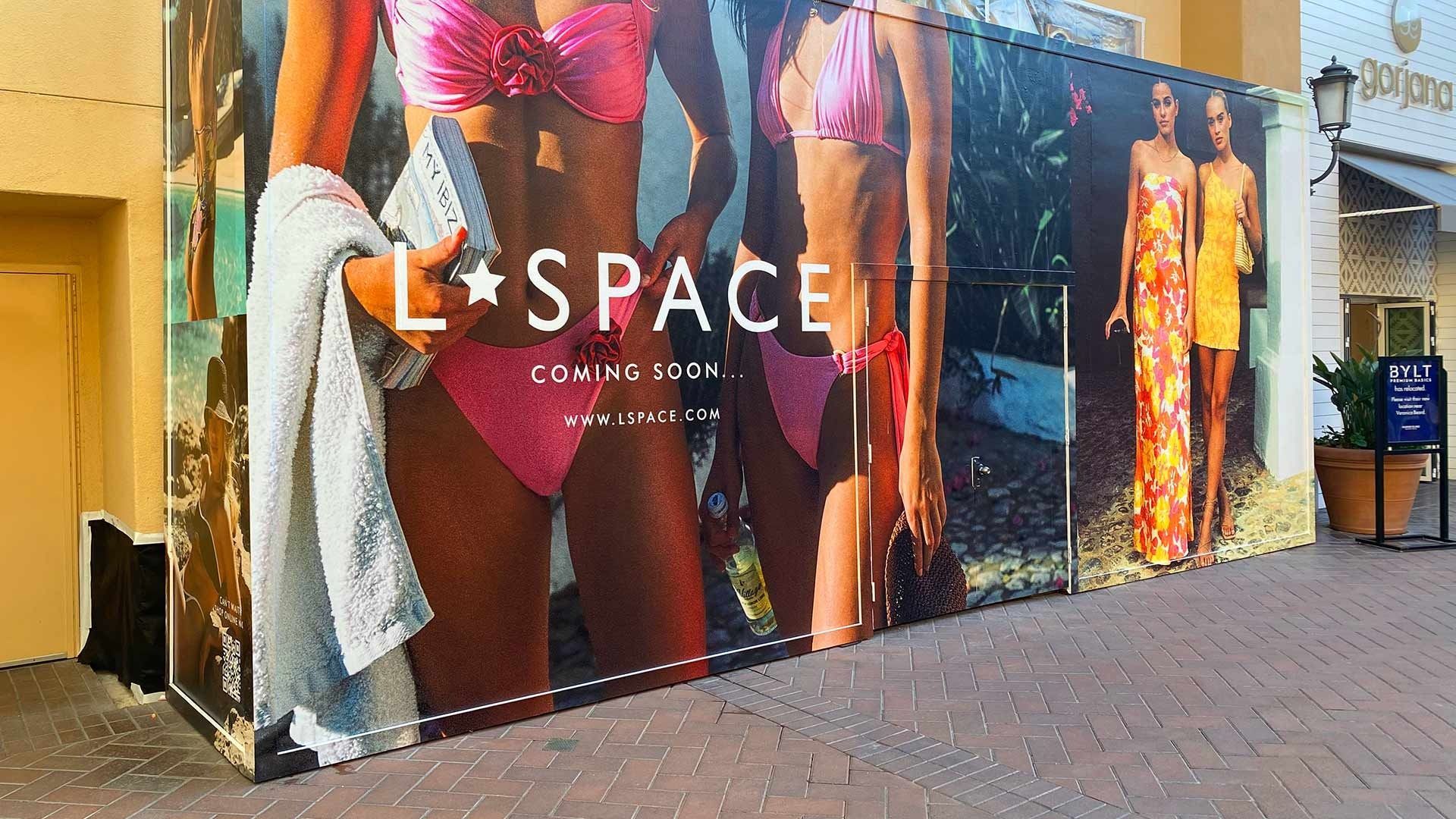 An L*SPACE store construction hoarding displaying promotional images of swimwear and clothing on a paved walkway.