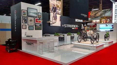 A trade show booth featuring a display of Stromer electric bicycles, white counters, and a photo collage on a black wall.