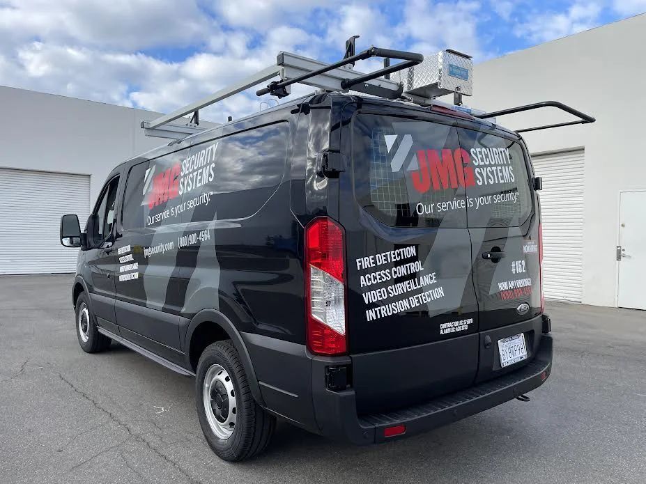 A black JMG Security Systems service van parked outside a commercial building with a roof ladder rack.