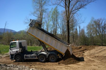 A white dump truck raising its bed to unload soil on a dirt field with trees and mountains in the background.