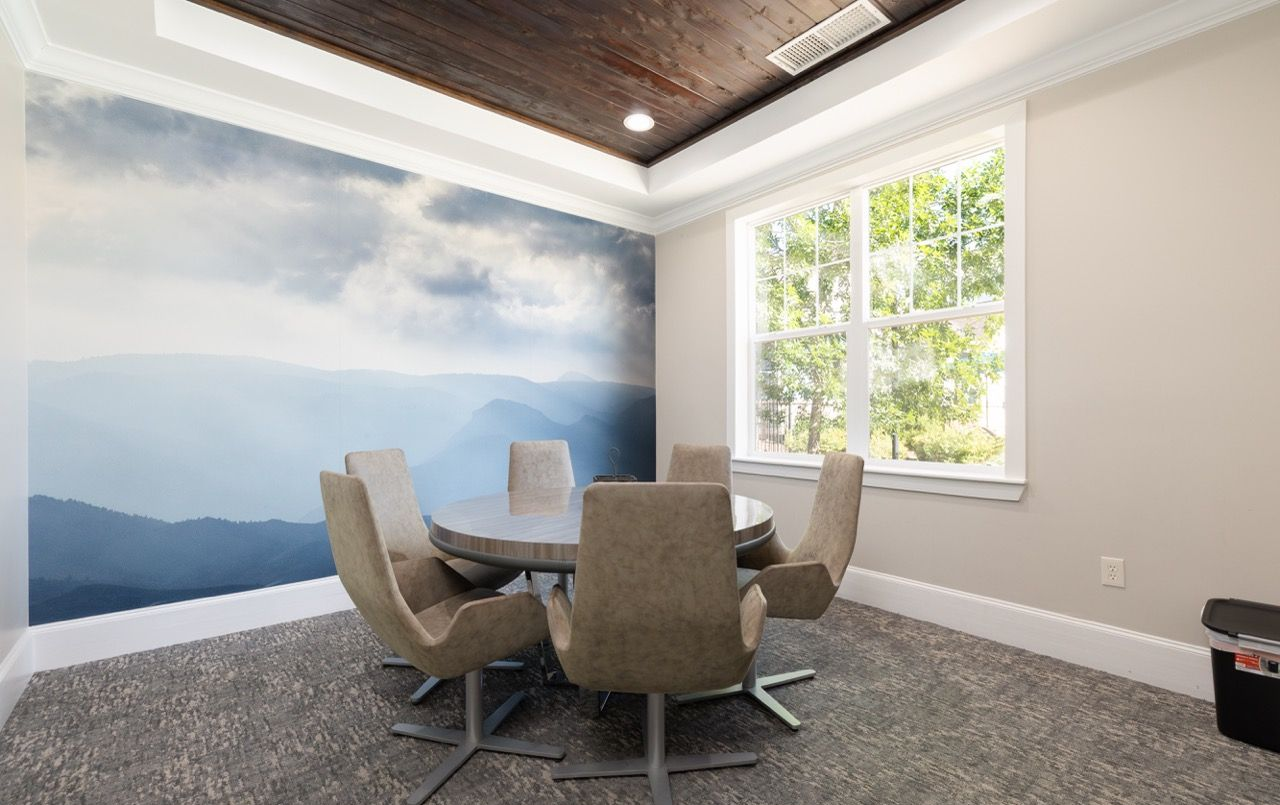 Indoor community meeting room with a round table, beige chairs, a large window, and a mountain mural.