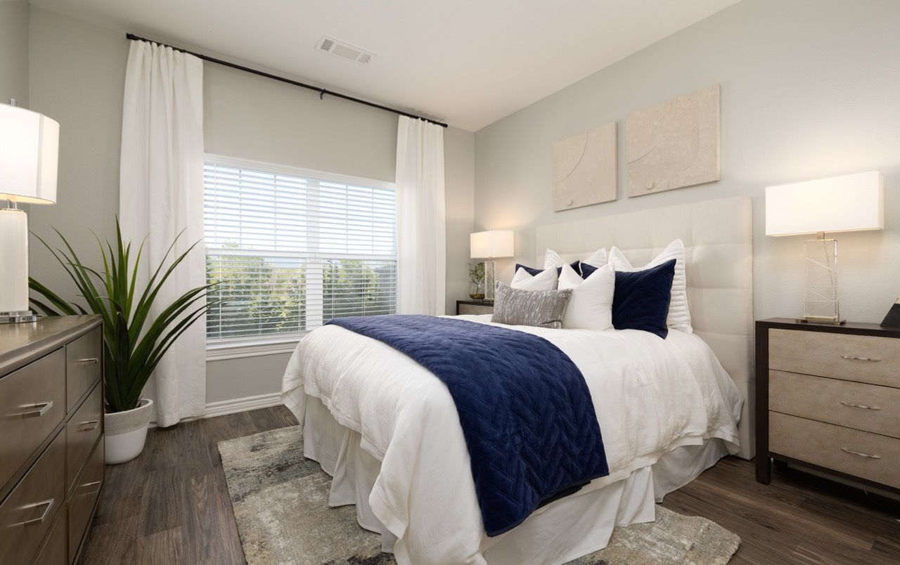 Modern furnished bedroom with white bedding, navy throw, nightstands, and a large window.