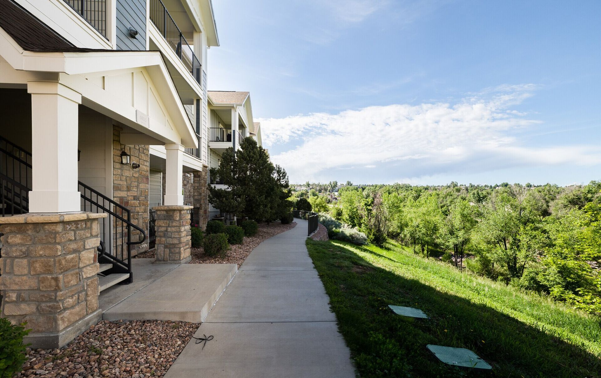 Apartment building exterior with sidewalk along a grassy slope, trees and a partly cloudy sky.
