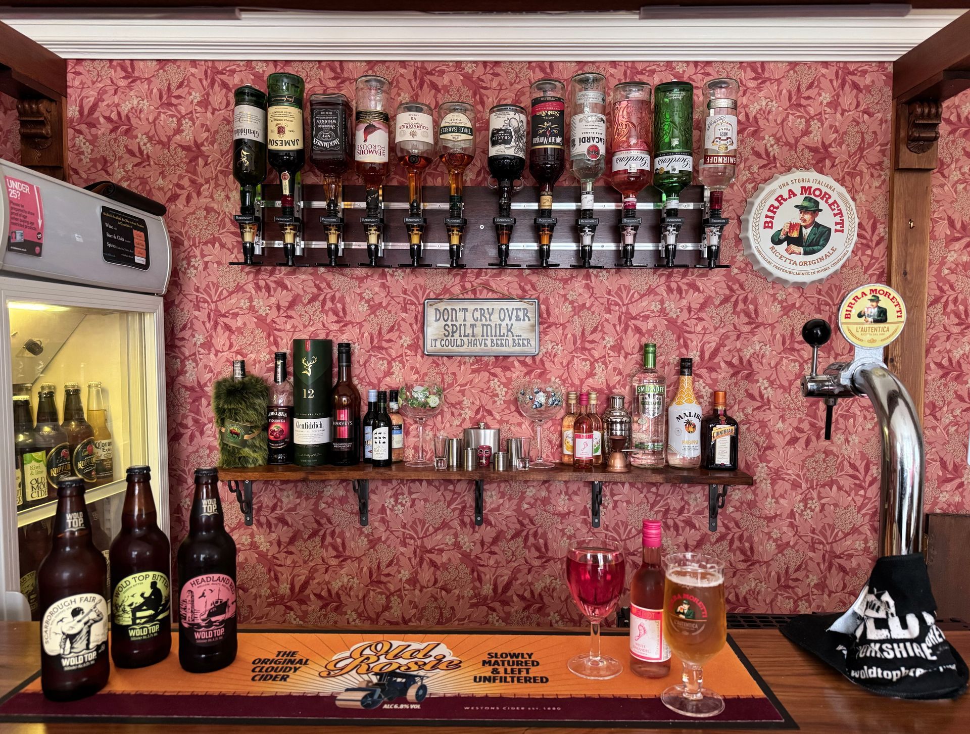 Bar with bottles, tap, drinks, and a small sign, with red floral wallpaper.