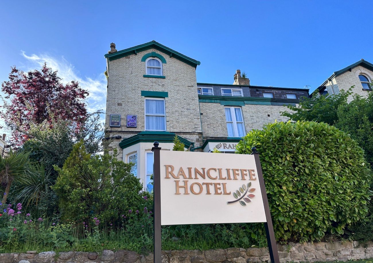 Raincliffe Hotel sign in front of a light brick building with green trim and a blue sky backdrop.