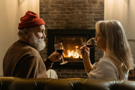 A man and a woman are sitting on a couch drinking wine in front of a fireplace.