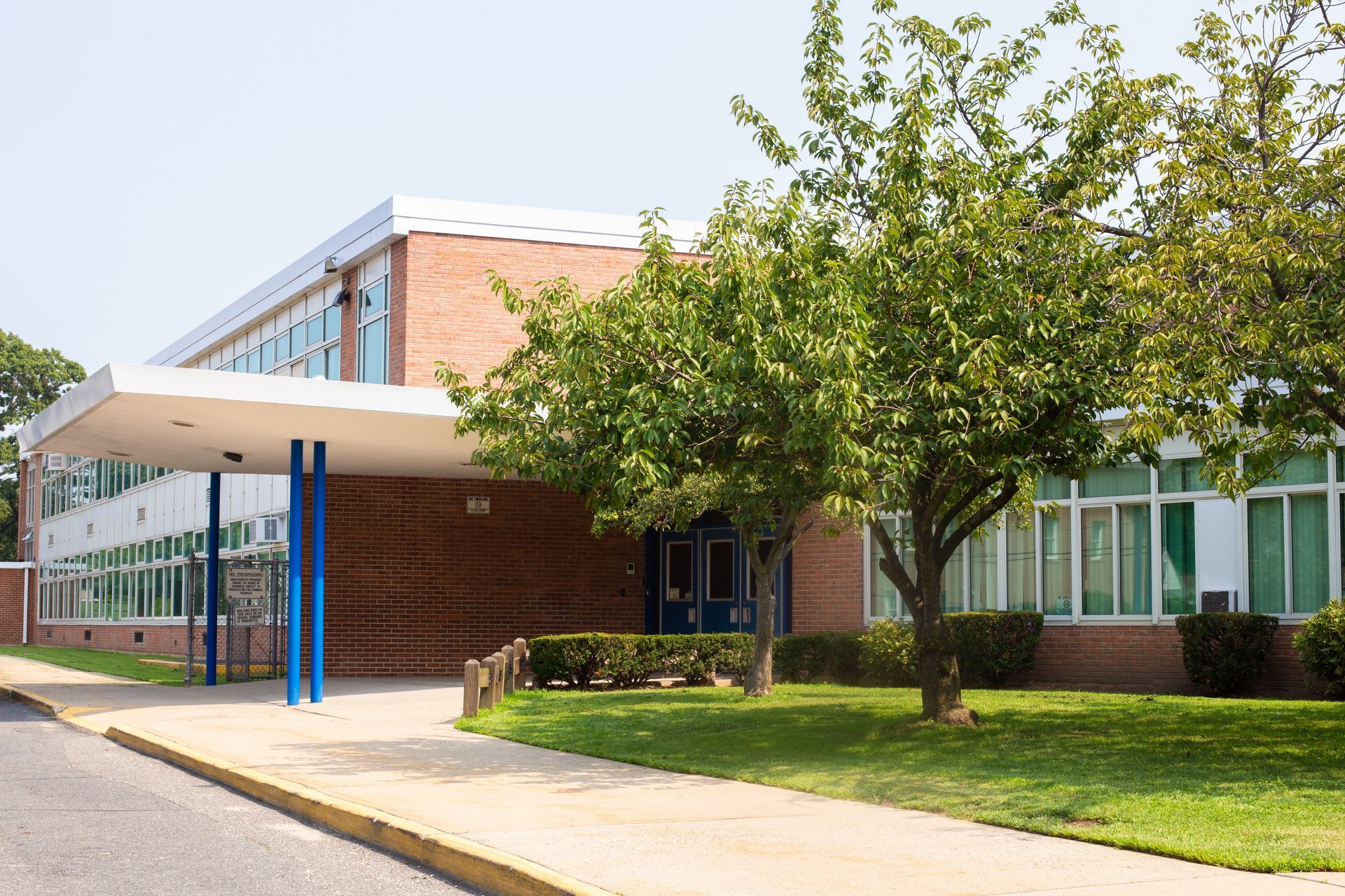 School building with brick facade, covered entrance, and trees in front on a sunny day.