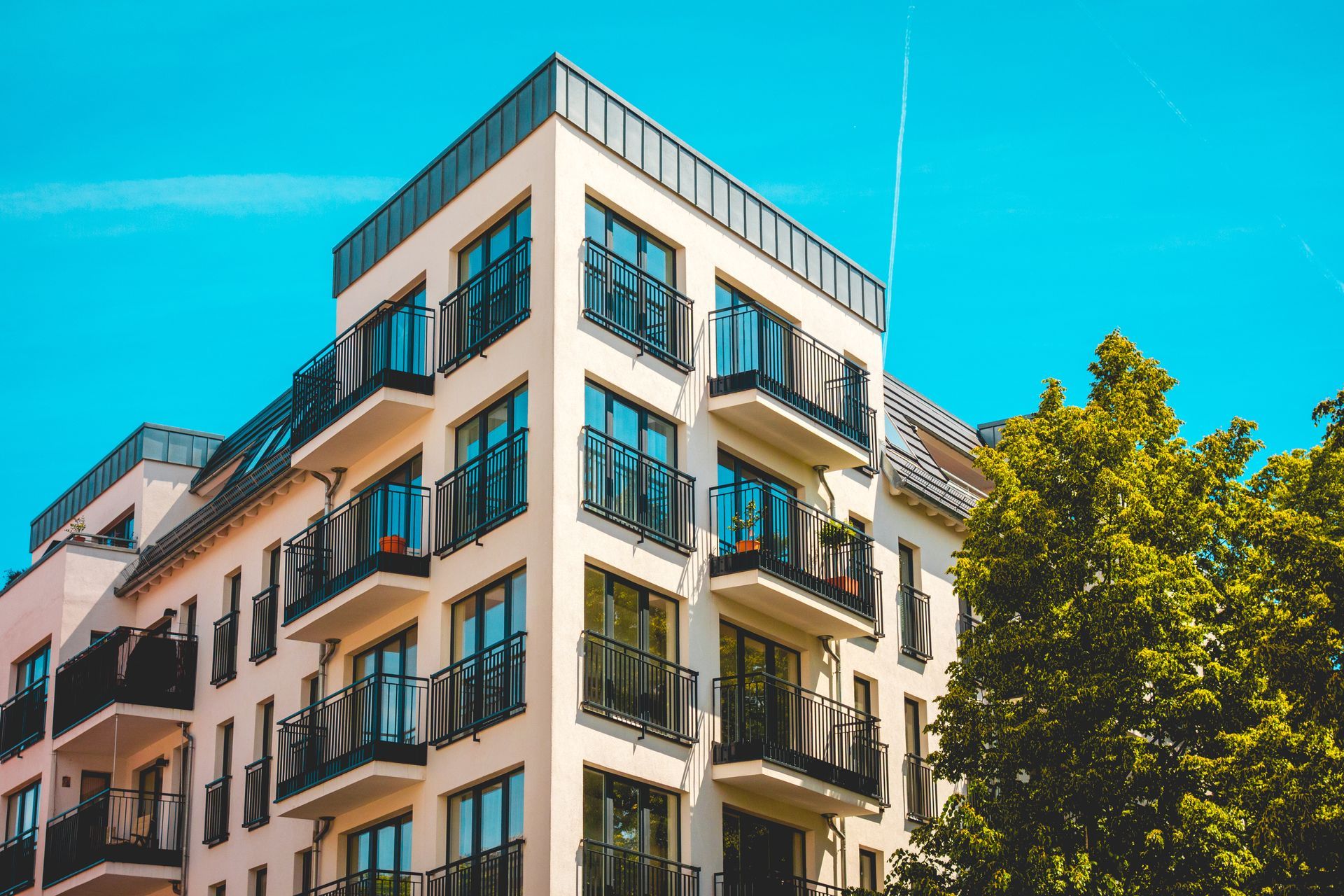 Modern, cream-colored apartment building with black balconies against a clear blue sky and lush green tree.
