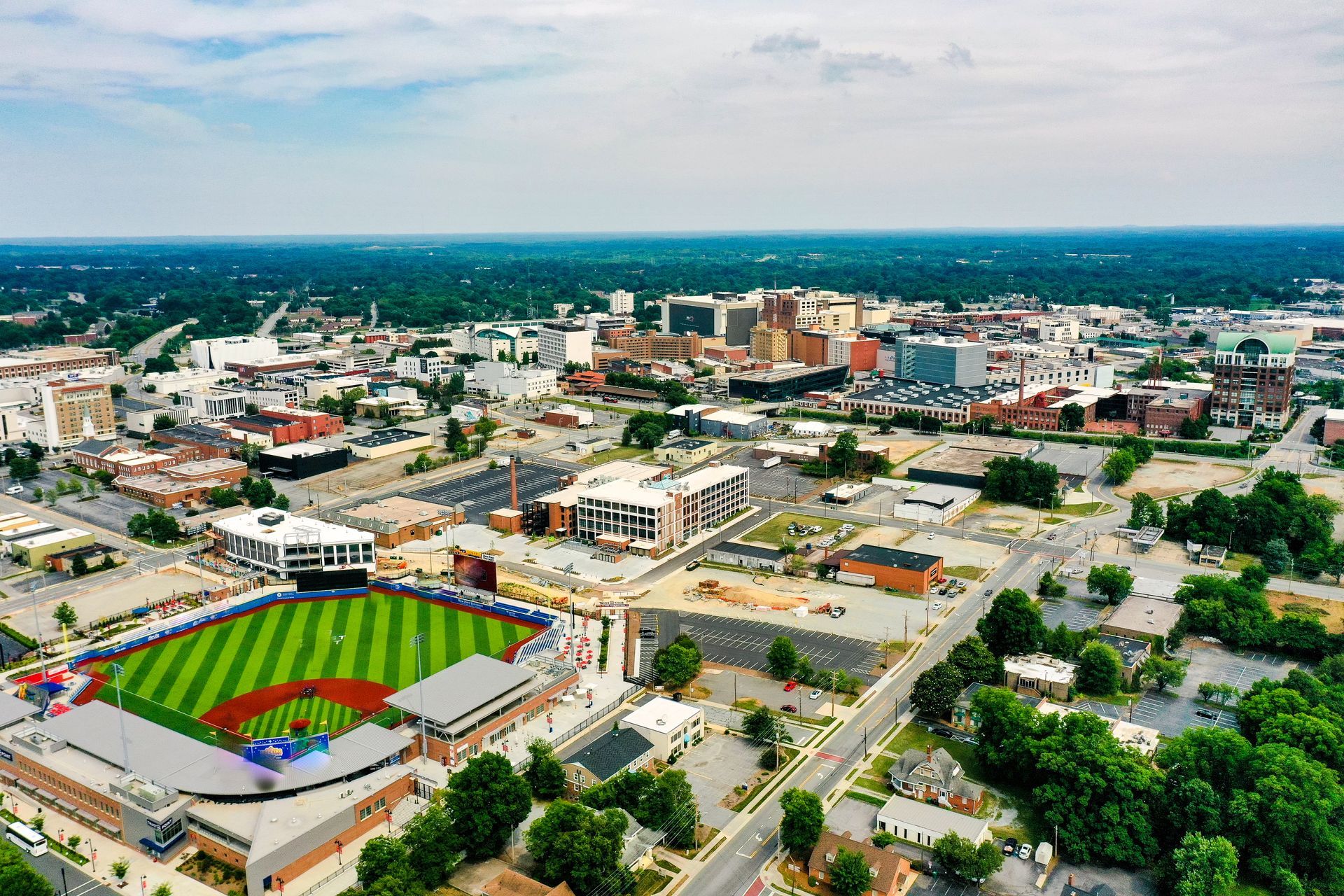 Aerial view of a city with a baseball field in the foreground; several buildings and green trees are visible.