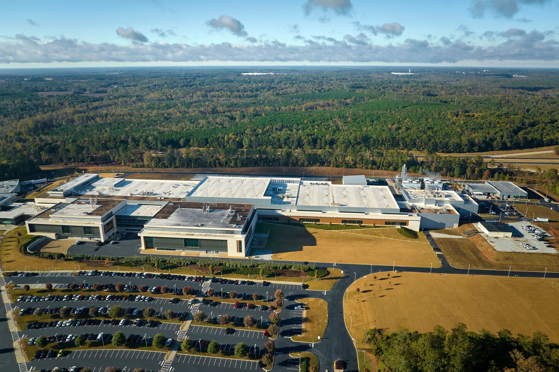 Aerial view of a large industrial complex with several buildings, surrounded by parking lots and a forest.