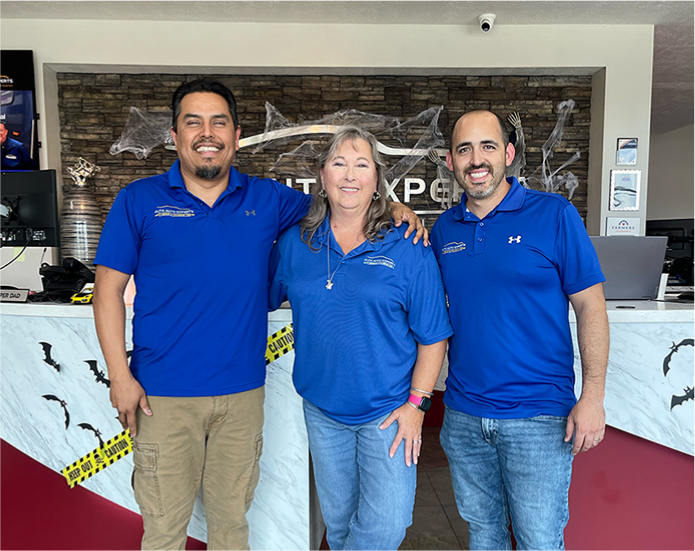 Three people in blue shirts are posing for a picture in front of a counter. | Elite Auto Experts