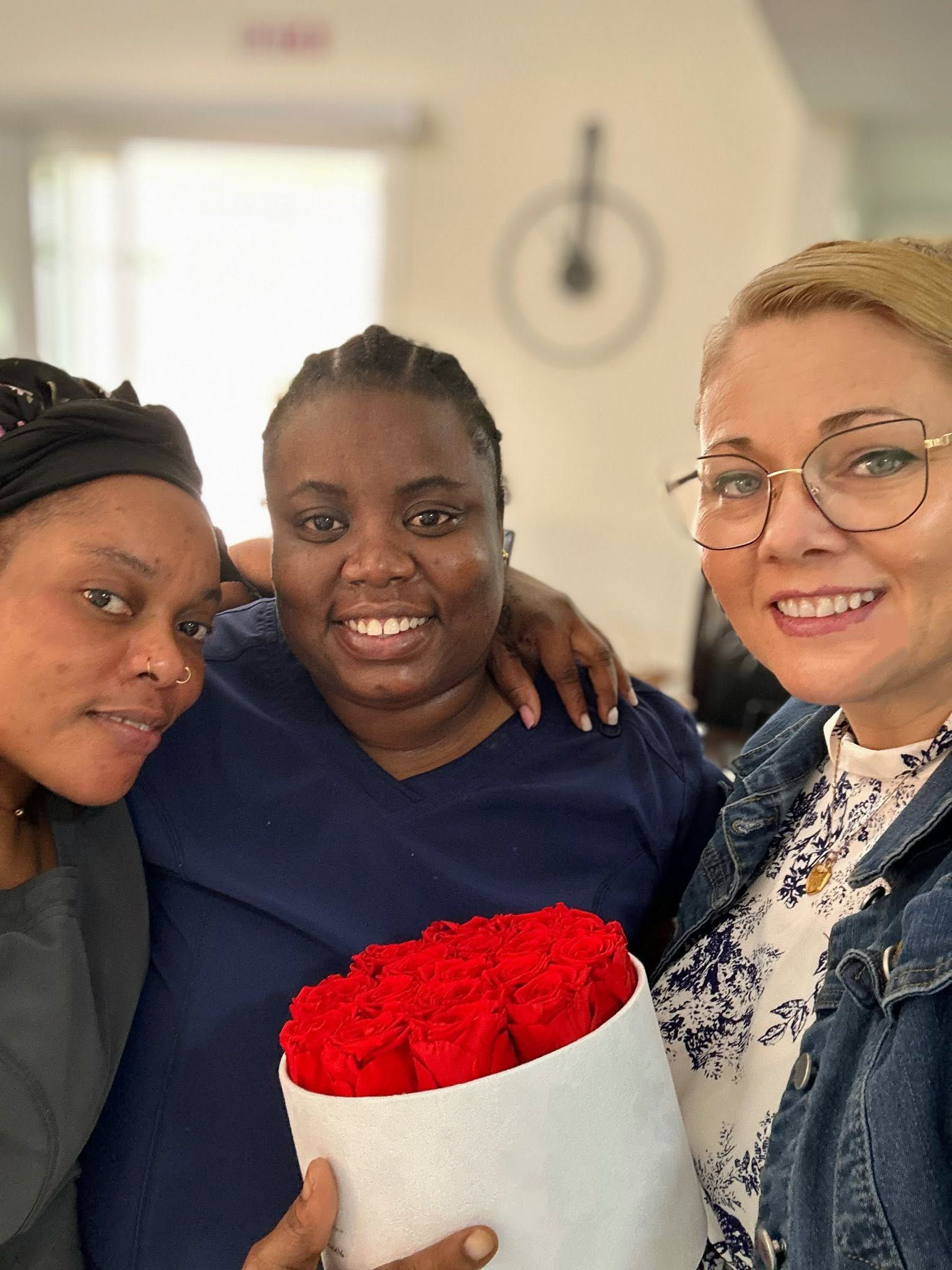 Three smiling people indoors, one holding a white vase of red flowers.