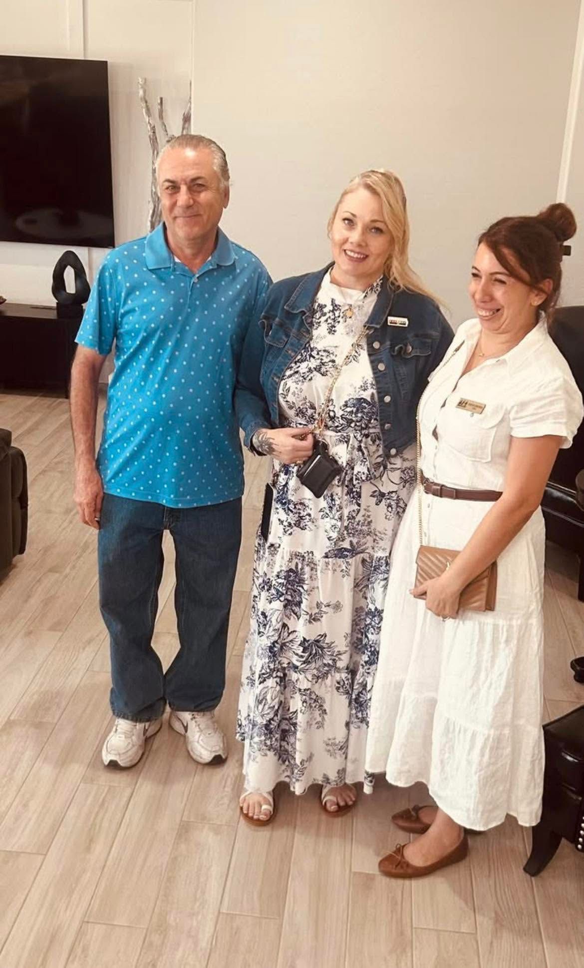 Three people posing indoors, smiling, with a man in blue and two women in white dresses