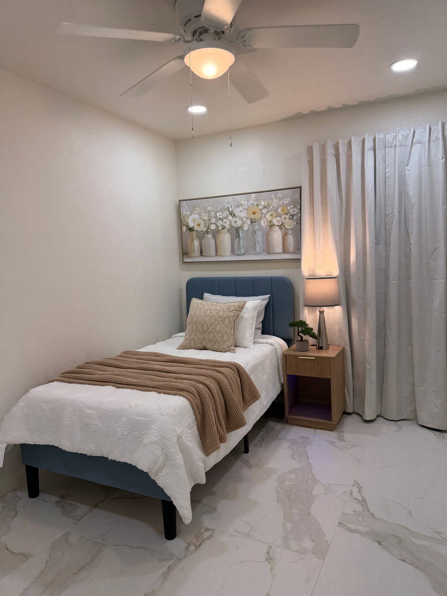 Small bedroom with a single bed, white curtains, ceiling fan, and bedside lamp on tiled floor.