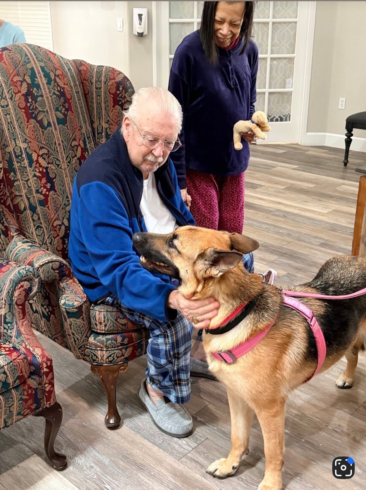 Man in a blue jacket pets a tan dog on a pink leash while a woman stands behind them indoors.
