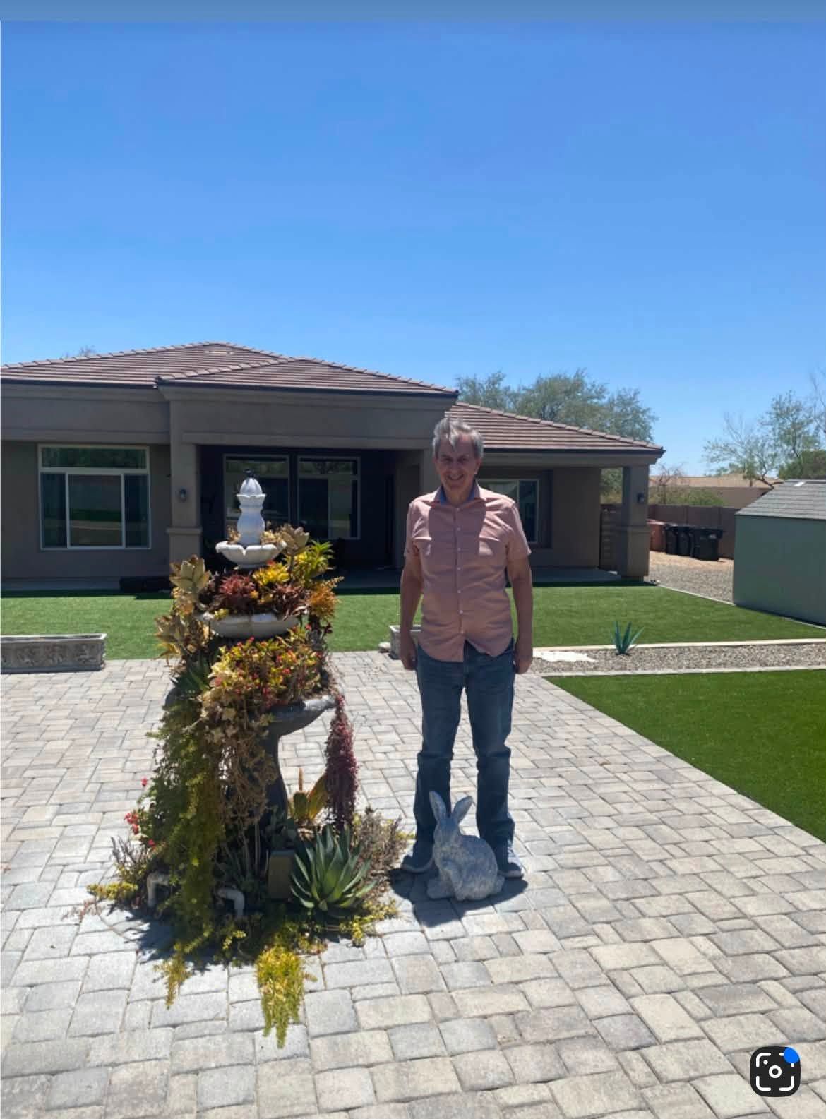 Person stands beside a floral arrangement on a sunny driveway in front of a house.