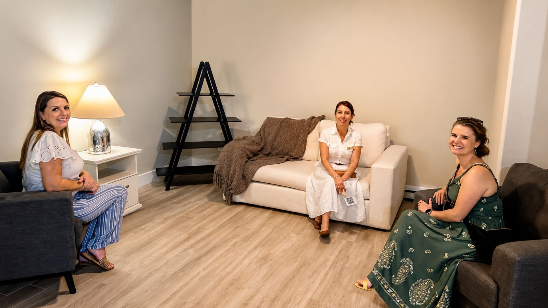 Three women seated in a bright living room, with a lamp, sofa, and folding rack in the background.