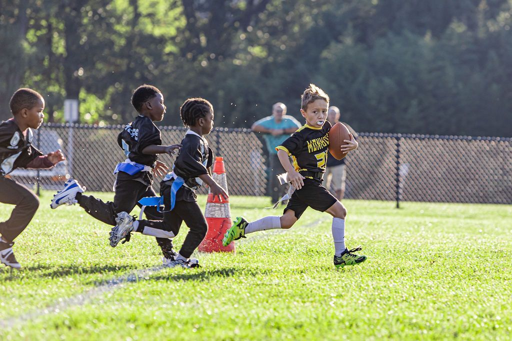Flag football game at night, a player in red jersey runs with the ball as others pursue on a green field.