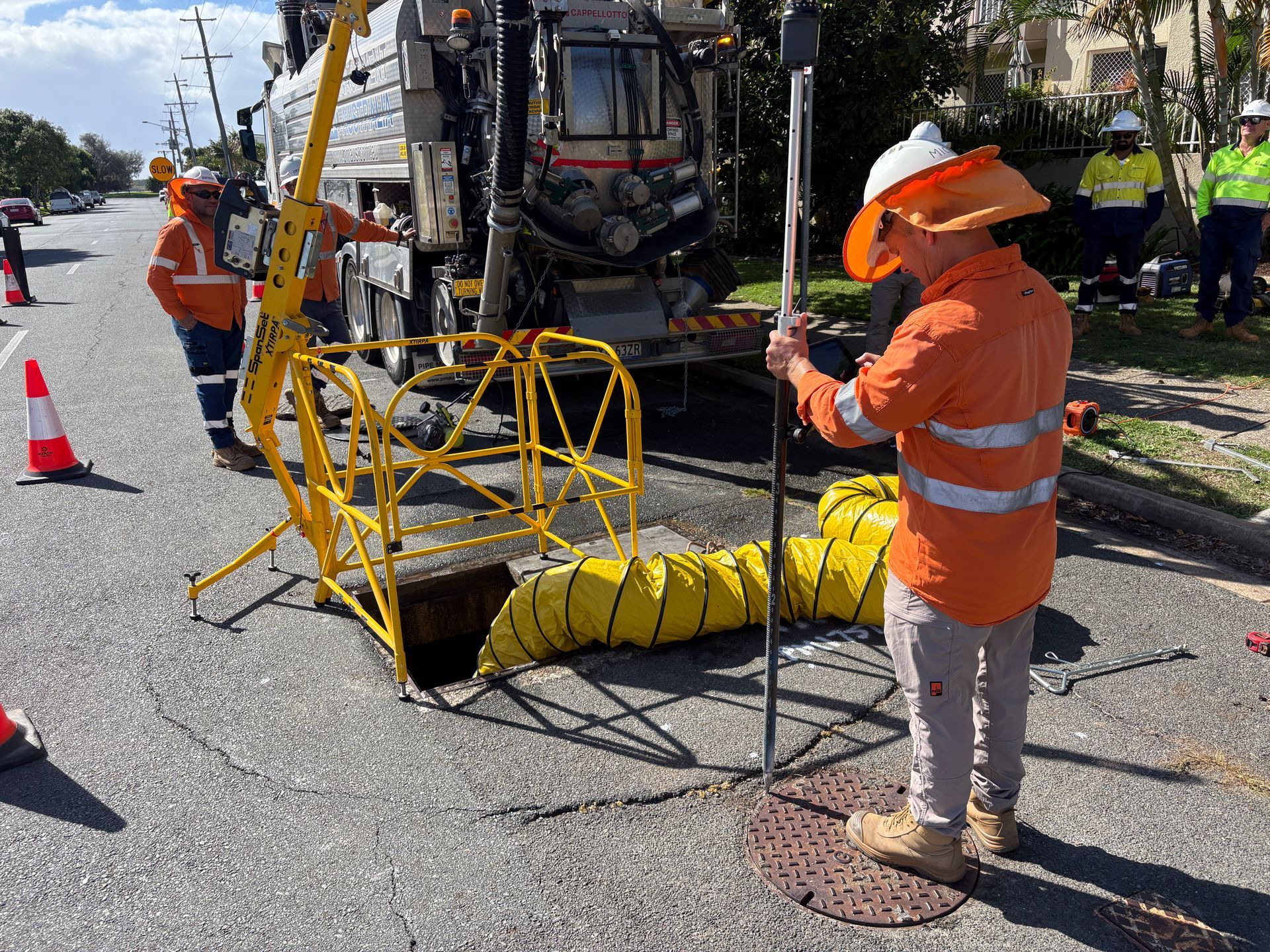Workers in high-visibility orange uniforms operate utility maintenance equipment at an open manhole on a paved street.