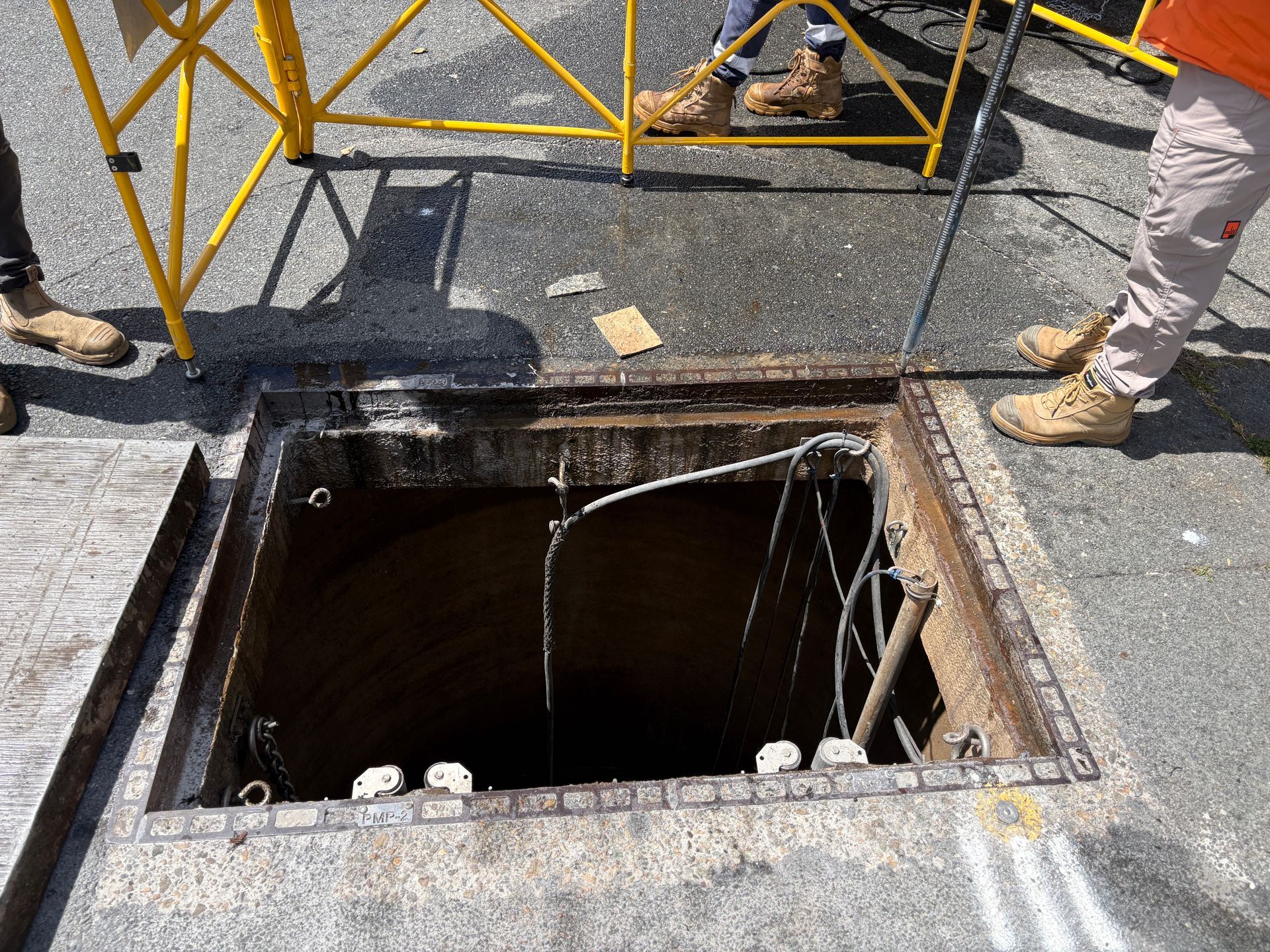 An open maintenance manhole in the pavement, surrounded by yellow safety barriers and the legs of workers in boots.