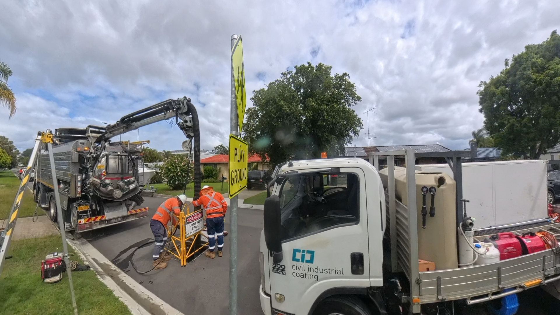 Utility workers in high-visibility gear work near a vacuum excavation truck and a white service vehicle on a suburban road.