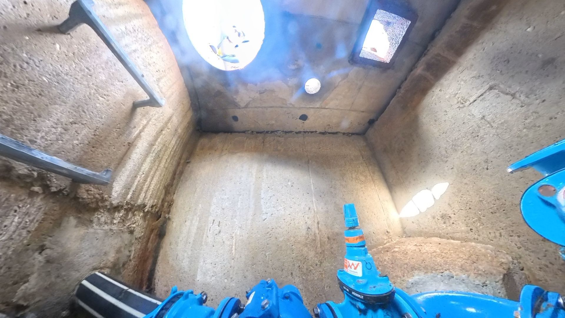 View from inside an underground concrete utility vault, showing blue industrial piping, valves, and a ladder on the wall.