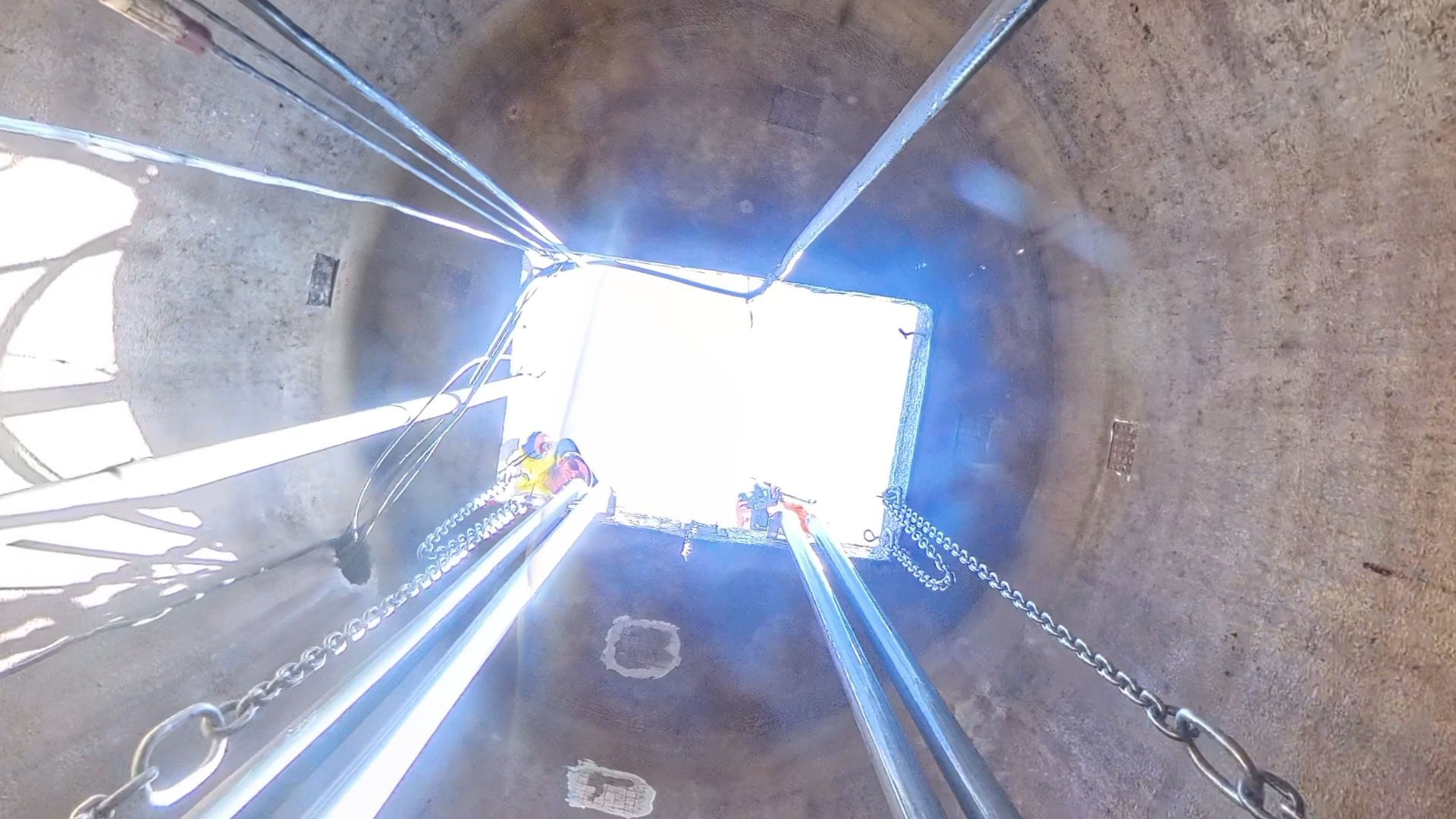 Low-angle view inside a concrete sewer pump station looking up at a bright rectangular opening with hanging chains and rods.