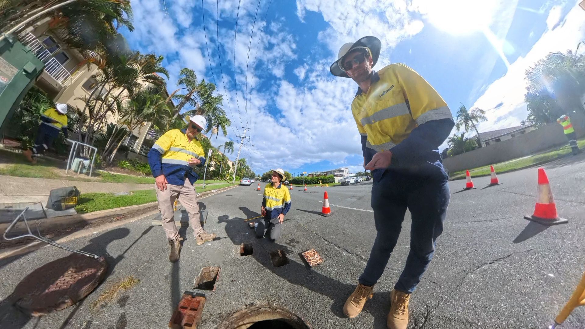 Three workers in high-visibility uniforms examine an open manhole on a sunny residential street with traffic cones.