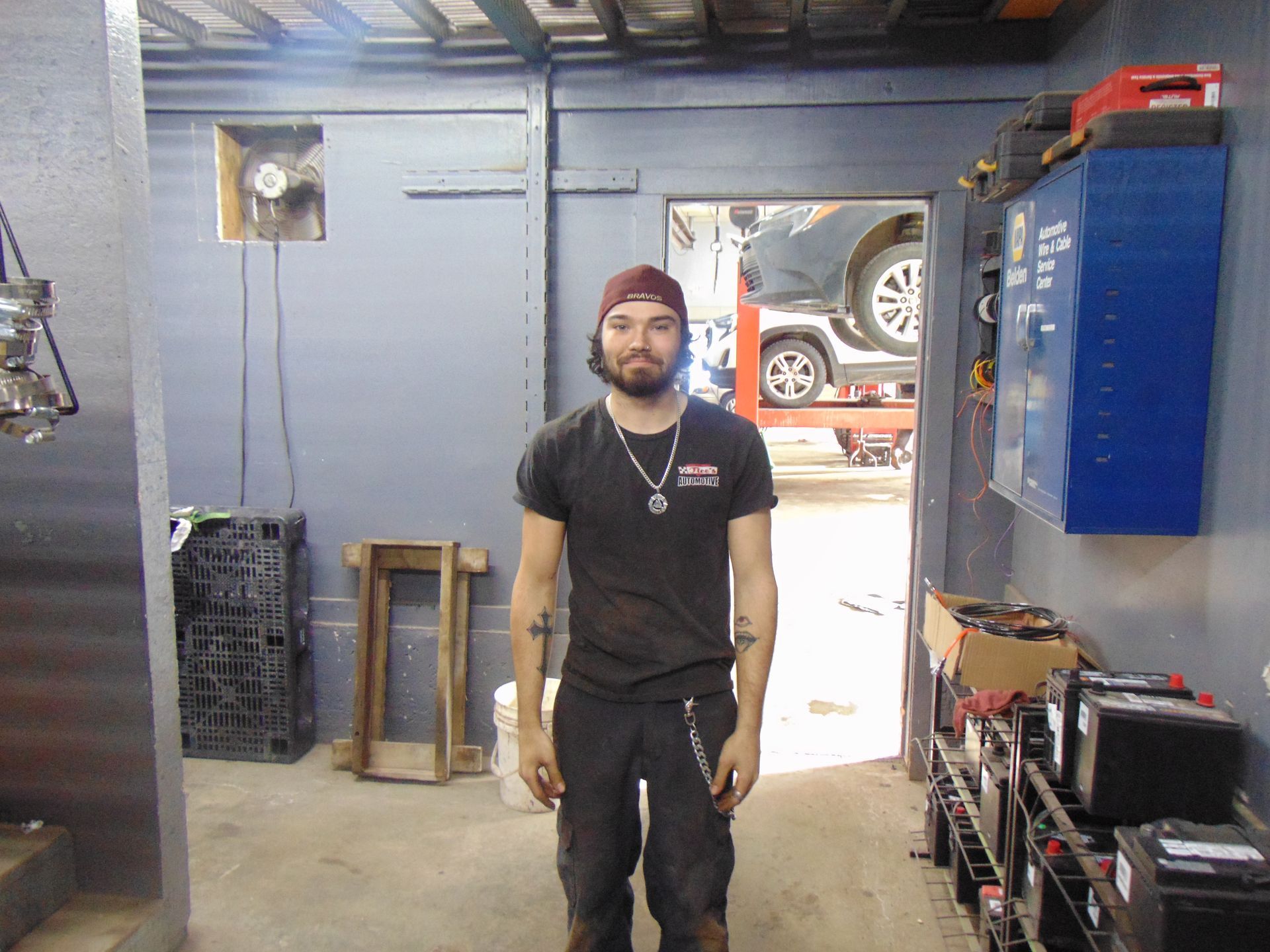 A person in a black t-shirt and work pants stands in an automotive repair shop with a car visible in the background.