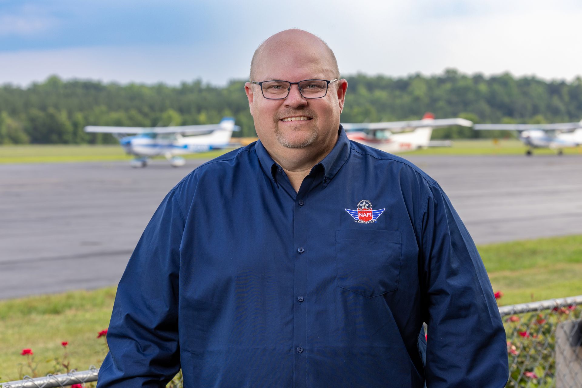 A man in a blue shirt is standing in front of a runway with planes in the background.