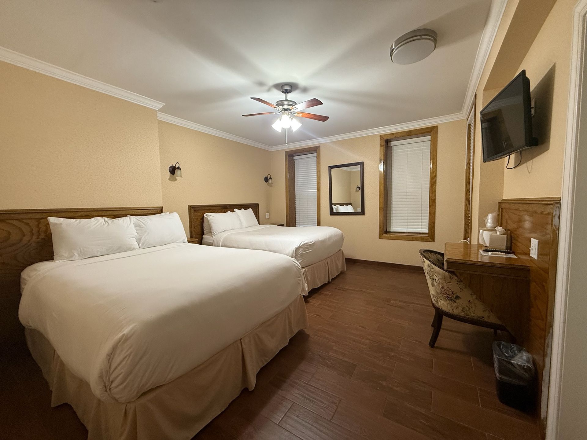 Hotel room with two queen-sized beds, wooden desk, and a mounted TV. Beige walls and wooden floor with a ceiling fan.