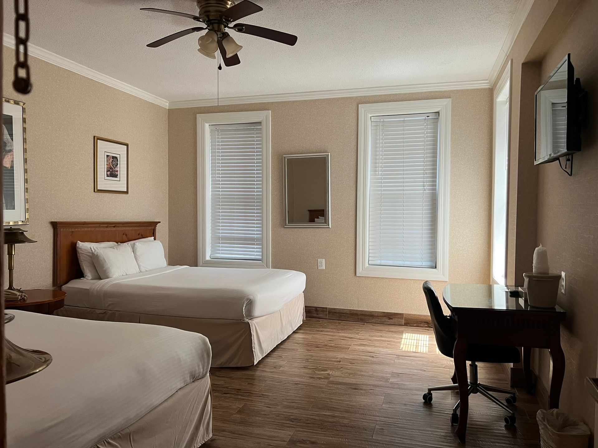 A hotel room with two beds, a desk, and two windows with blinds. The room has beige walls and a ceiling fan.