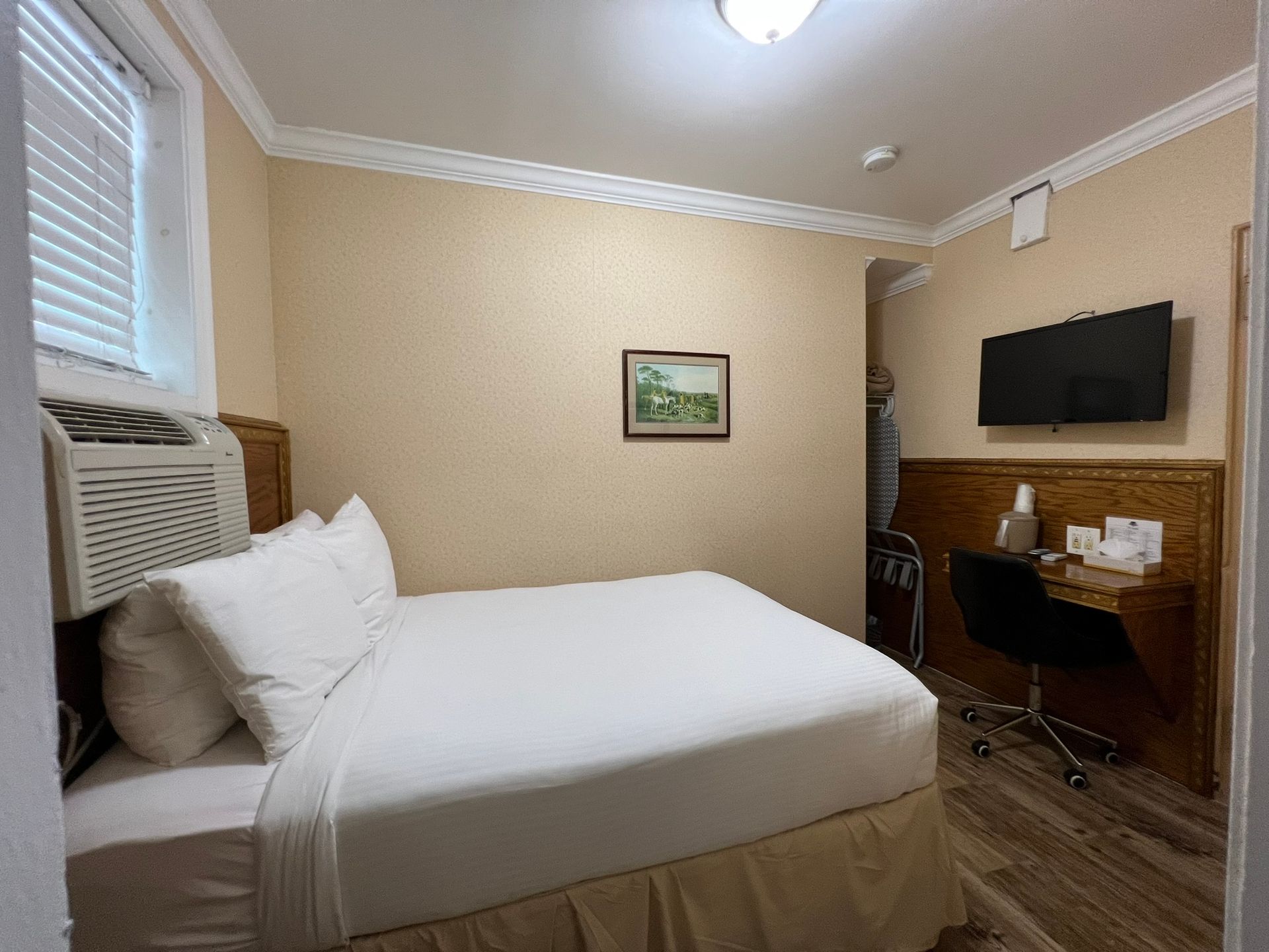 A small hotel room with a bed, desk, and TV. Beige walls and trim, a window with blinds, and a painting hang on the walls.