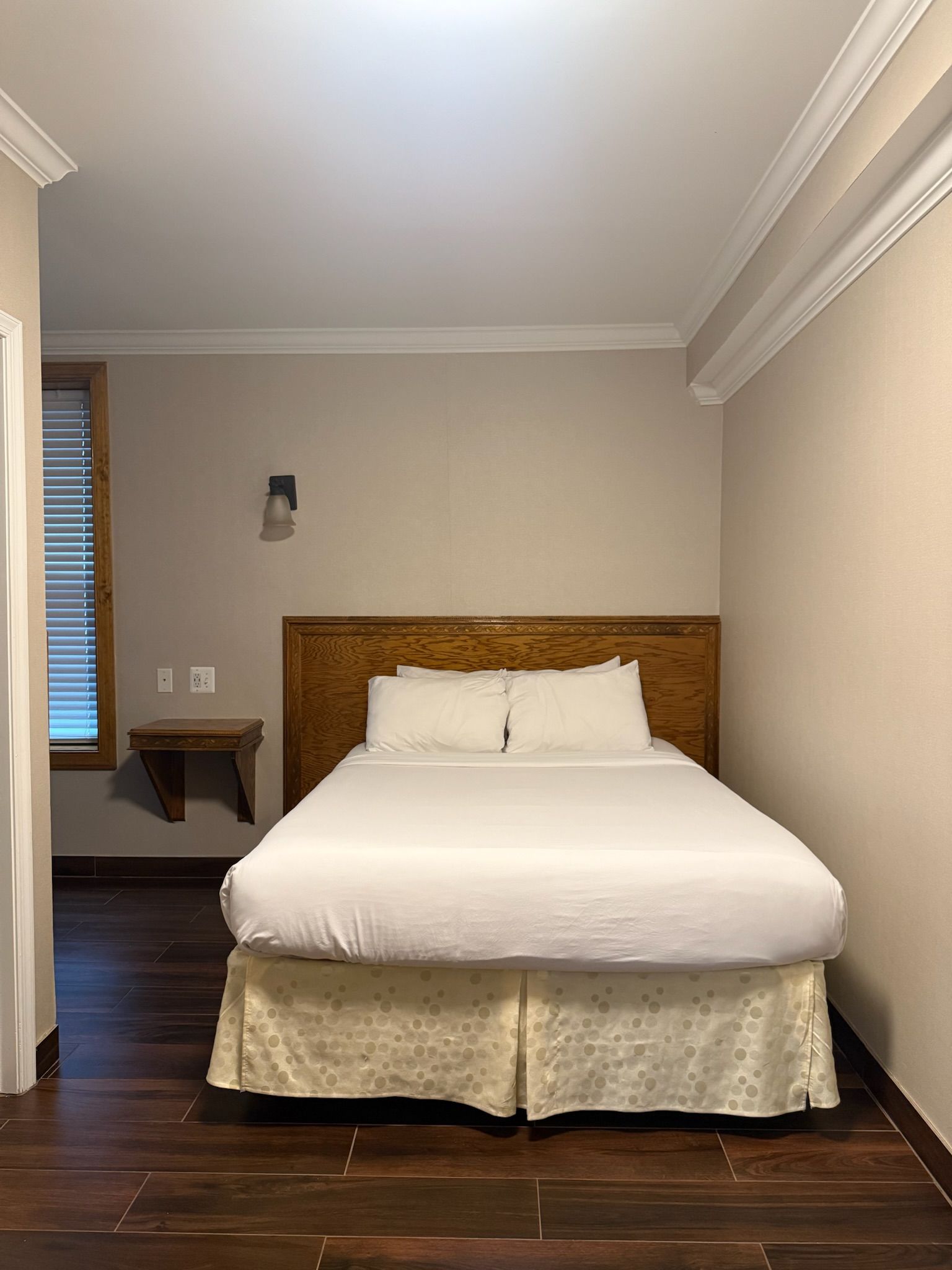 A hotel bedroom with a made bed, wooden headboard, and dark wood floor. There is a window and small shelf on the left side.