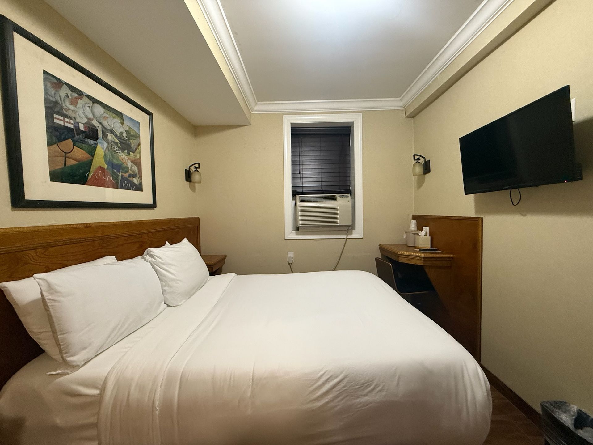 A hotel room with a bed, TV, and window. The room is painted beige and has a framed picture on the wall.