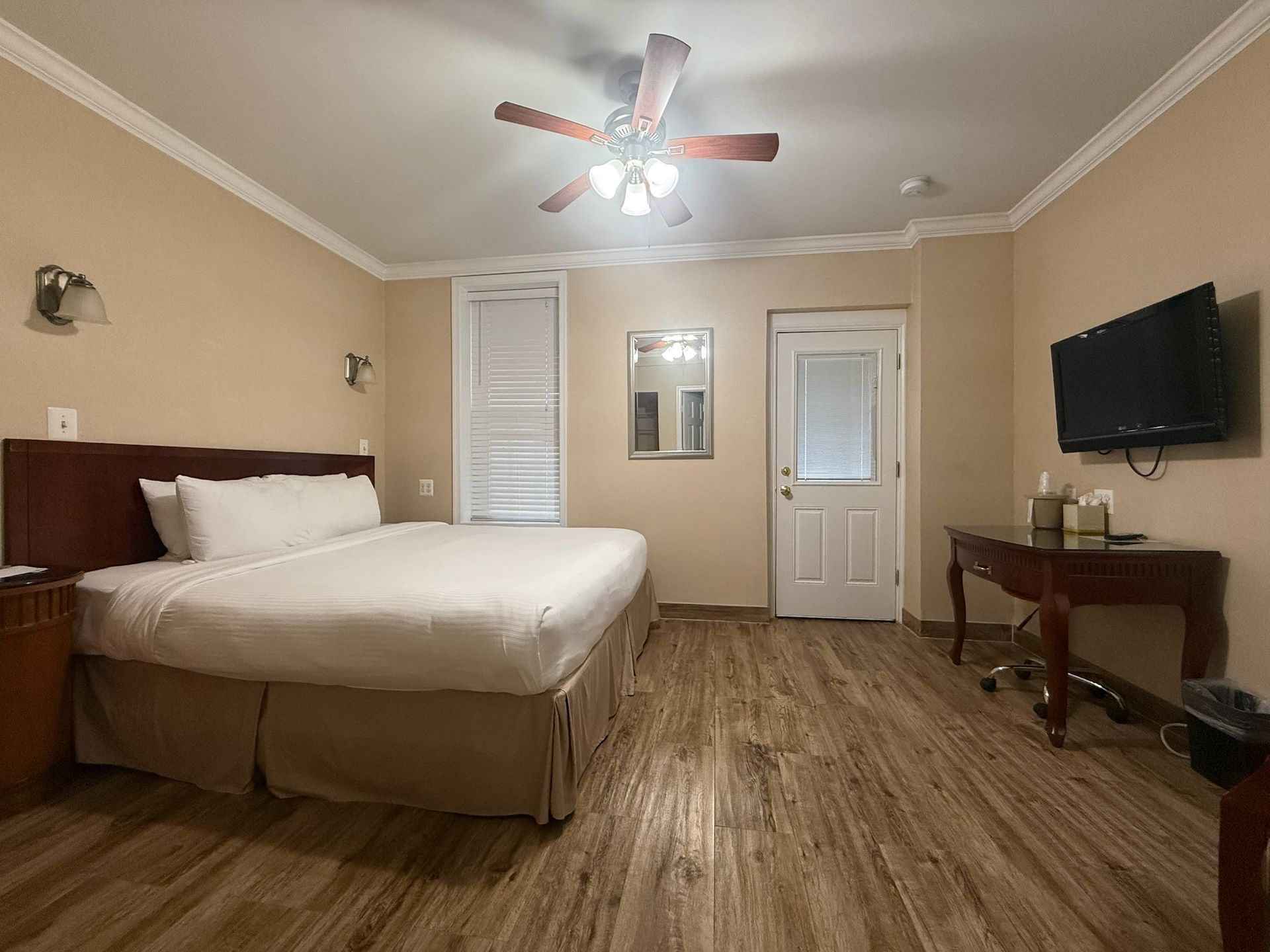 A hotel room with a king-size bed, desk, television, and a door leading outside. Wooden floors and light-colored walls.