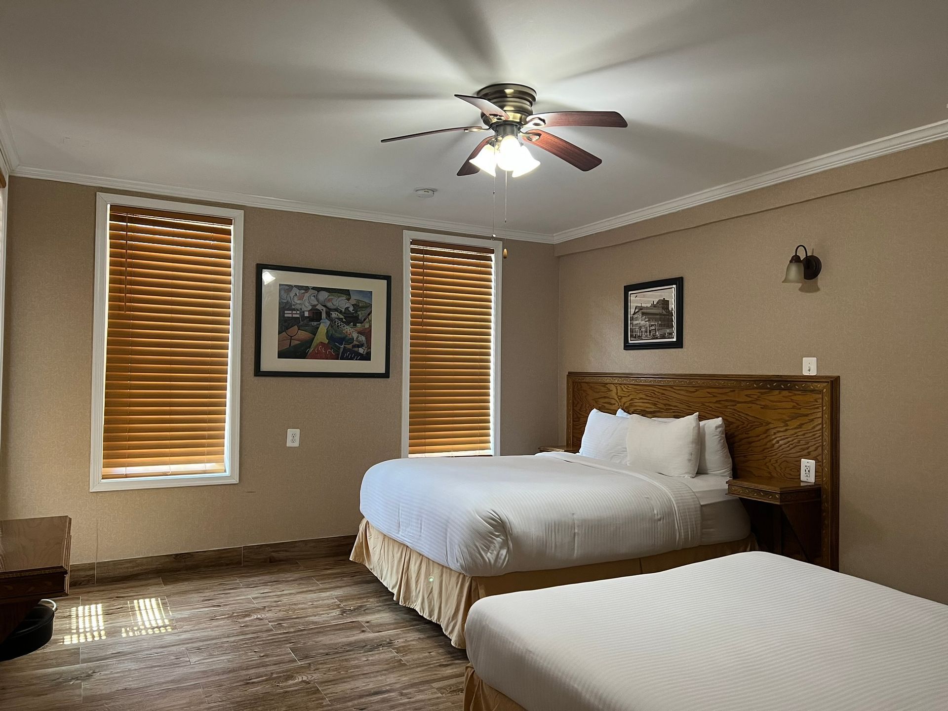 A hotel room with two queen beds, two windows with blinds, and artwork on the wall. Tan walls and ceiling with a ceiling fan.