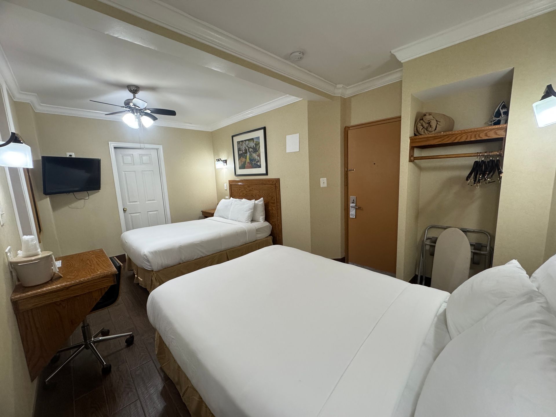 A hotel room with two beds, a TV, desk, and closet. The walls are beige, and the beds have white linens.