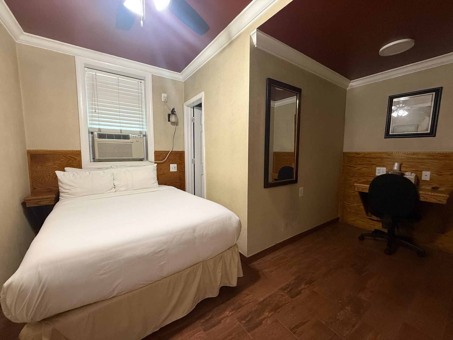 Small hotel room with a bed, desk, and a mirror. The room is illuminated by a ceiling fan light fixture.