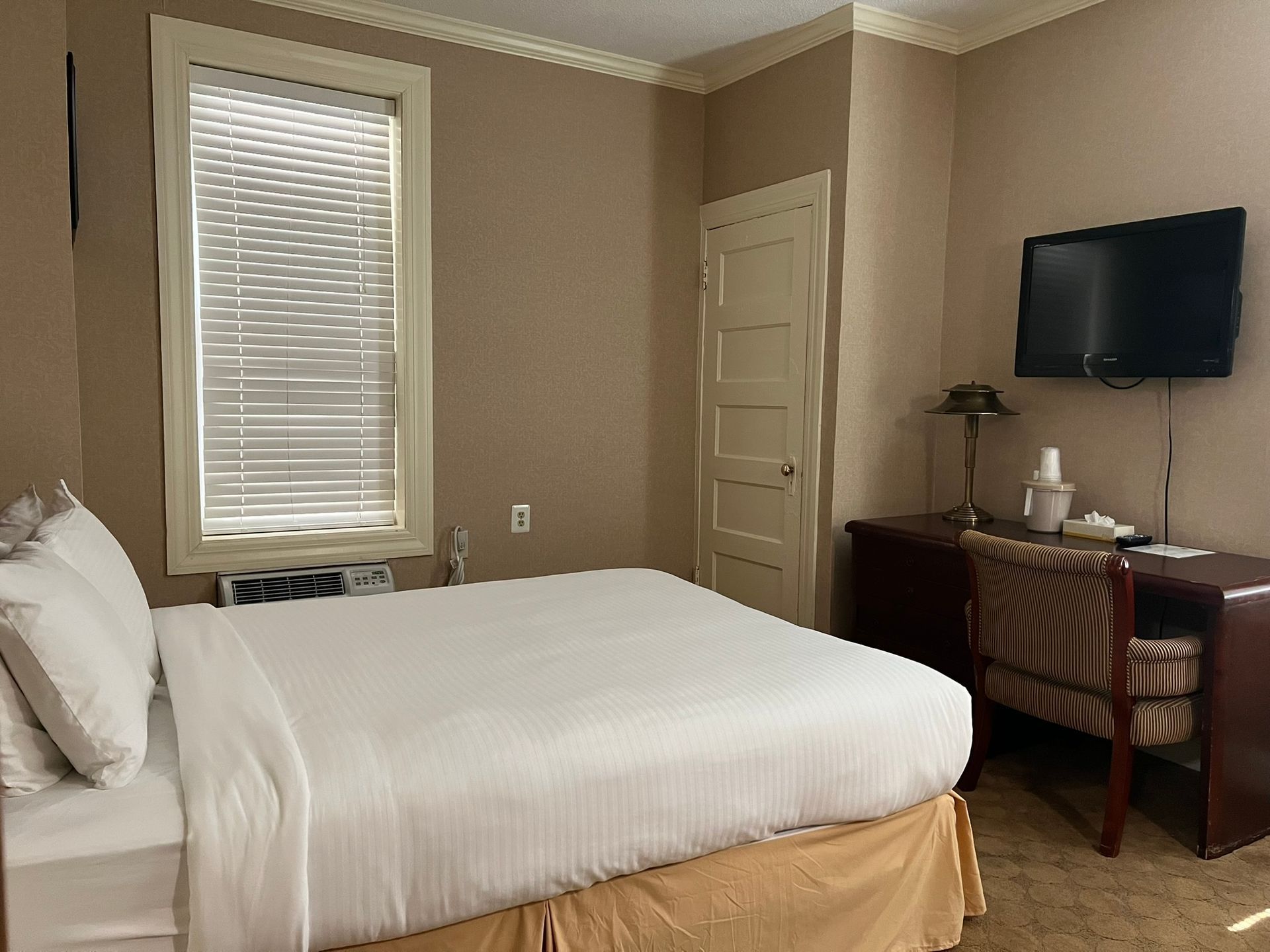 A hotel room with a bed, desk, TV, and window with blinds. The walls are tan, and the bed has white bedding.