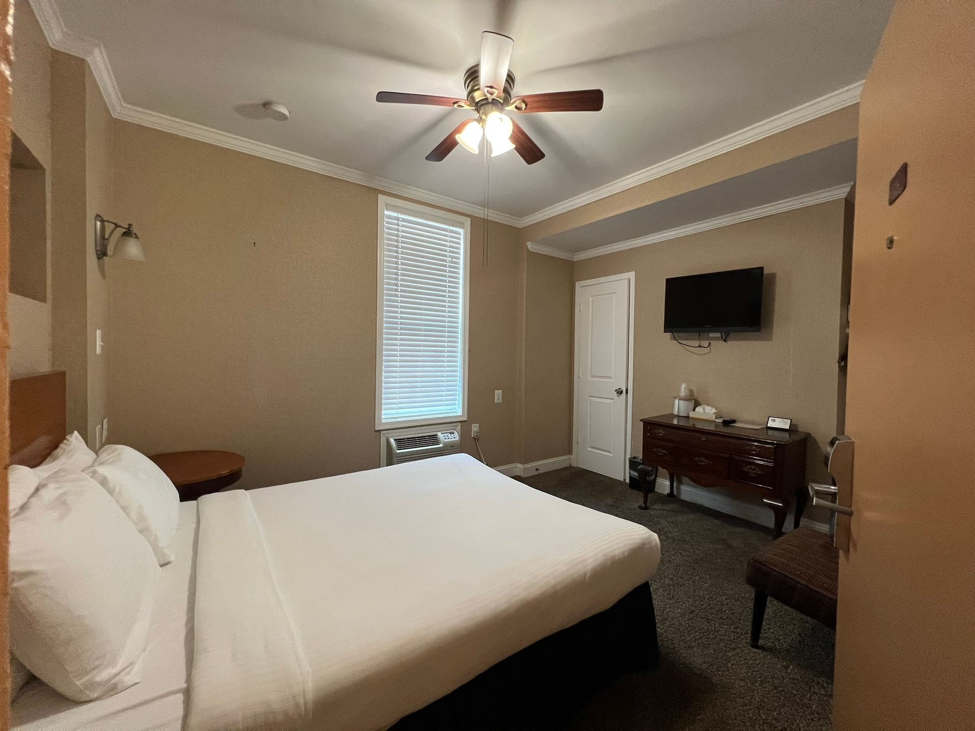 A hotel room with a bed, TV, dresser, and window. Beige walls, a brown ceiling fan, and white bedding are visible.