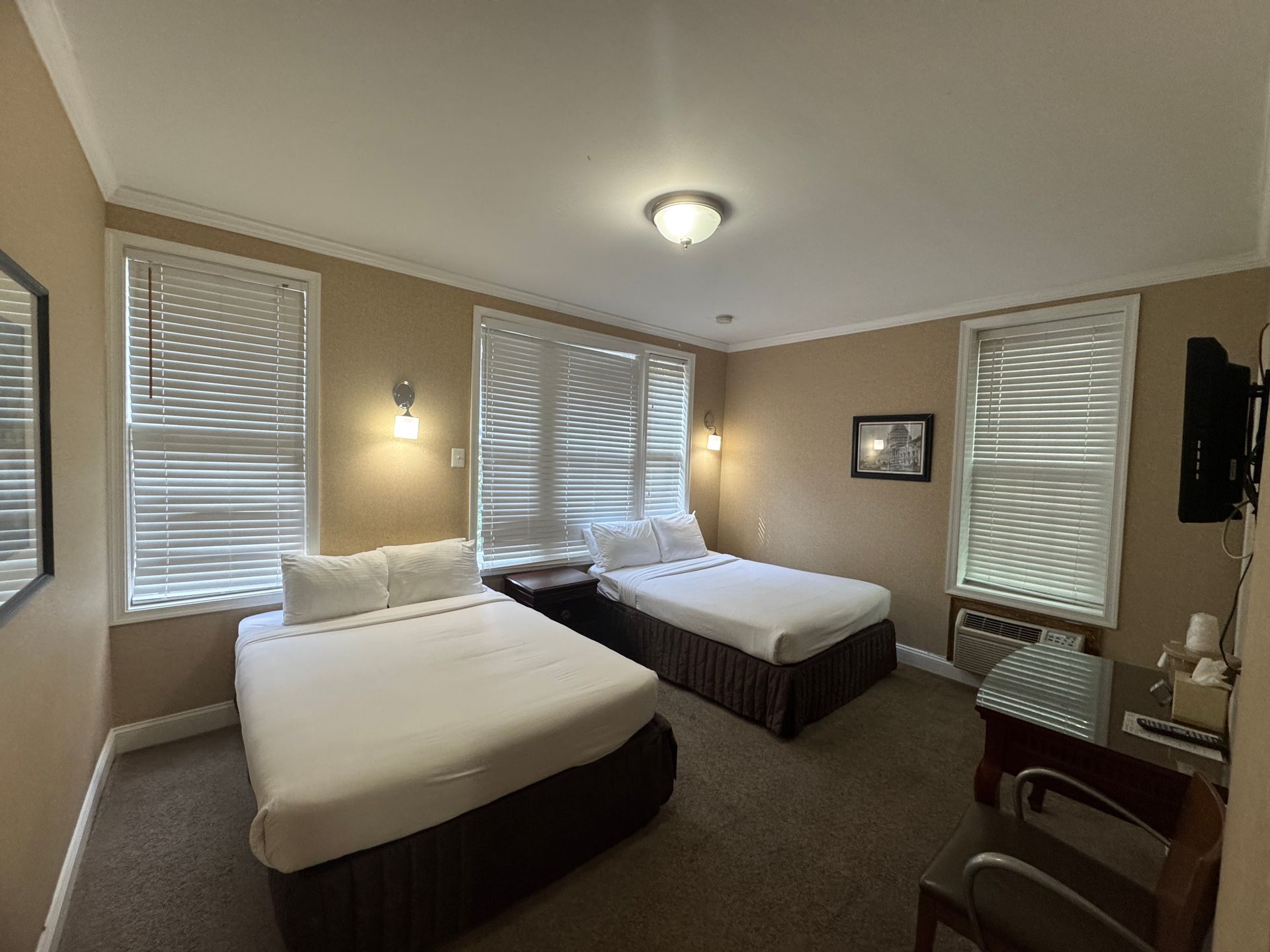 A hotel room with two beds, two windows, and a desk. The walls are beige and the carpet is brown.