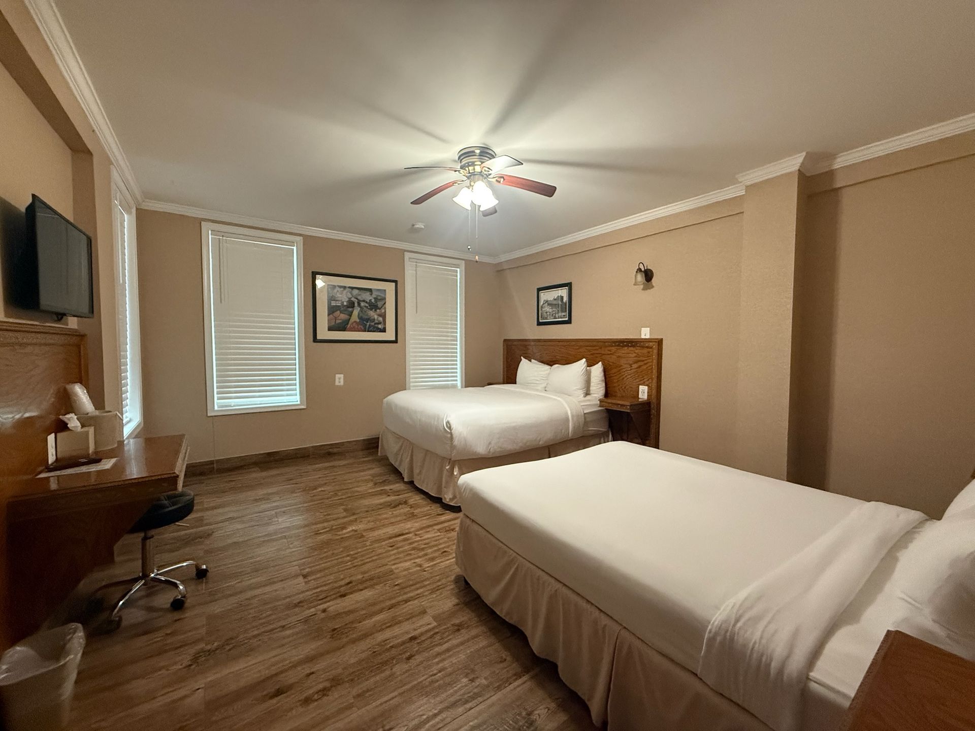 A hotel room with two beds, a desk, and a mounted TV. The room has wooden floors, beige walls, and is well-lit.